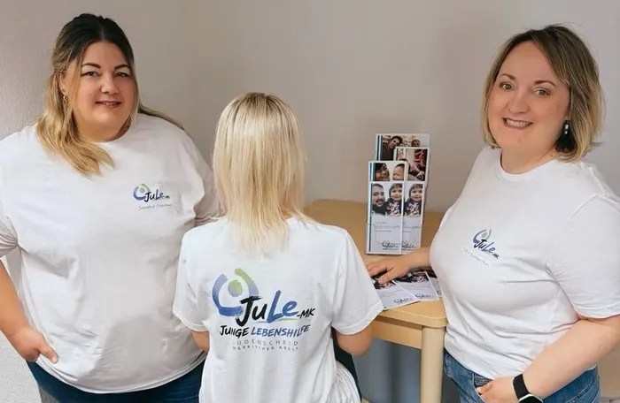 Drei Frauen in weißen T-Shirts mit dem Logo von „JuLe-MK Junge Lebenshilfe“ stehen an einem Tisch mit Informationsmaterial.