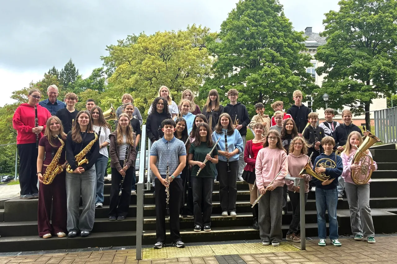 Eine Gruppe von etwa 25 Jugendlichen steht auf einer breiten Treppe vor Bäumen und Gebäuden. Viele halten Blasinstrumente wie Klarinetten, Saxophone, Trompeten und Posaunen.