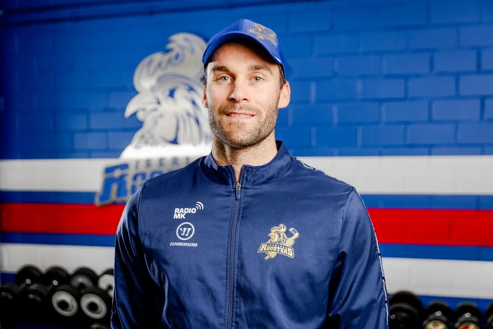 Justin Scott, neuer Spieler der Iserlohn Roosters, trägt Trainingskleidung mit dem Vereinslogo und lächelt in die Kamera.