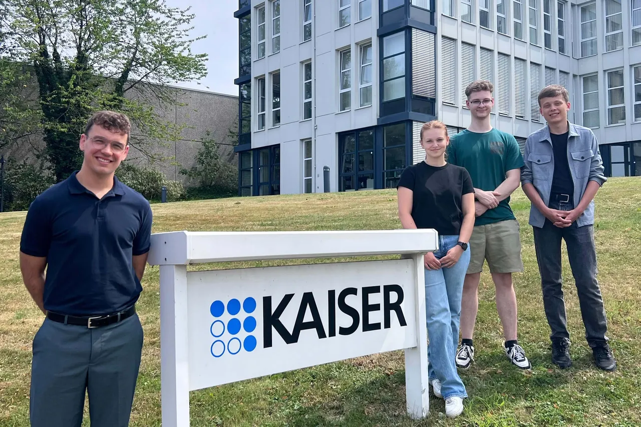 Vier Auszubildende stehen vor einem weißen Schild mit dem blauen „KAISER“-Logo vor einem modernen Gebäude.