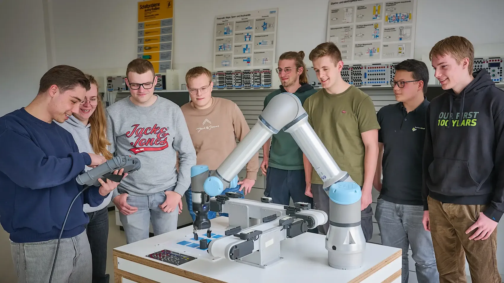 Eine Gruppe junger Menschen steht um einen Roboterarm in einem Werkraum.