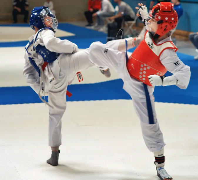 Zwei Taekwondo-Athleten in voller Schutzausrüstung kämpfen auf einer blauen Matte. Der Athlet im roten Anzug führt einen Tritt aus, während der Athlet im blauen Anzug ausweicht.