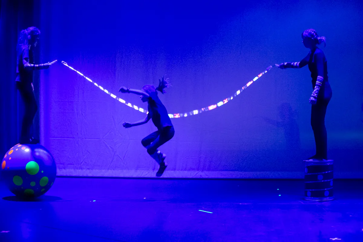 Drei Artisten unter blauem Licht: Eine Person springt, während zwei andere ein leuchtendes Seil halten. Links steht ein bunter Ball.