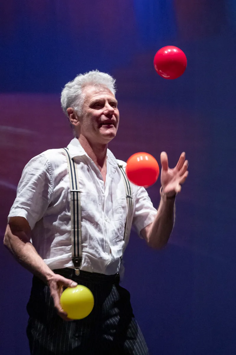Ein älterer Mann mit weißen Haaren jongliert mit drei Bällen auf einer Bühne. Er trägt ein weißes Hemd mit Hosenträgern und ein gestreiftes Jackett.