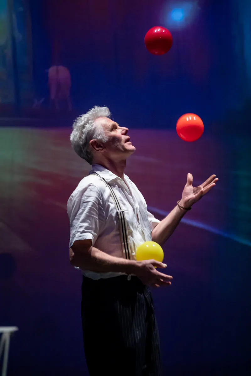 Ein älterer Mann mit grauen Haaren jongliert mit drei Bällen auf einer Bühne vor einem verschwommenen Hintergrund.