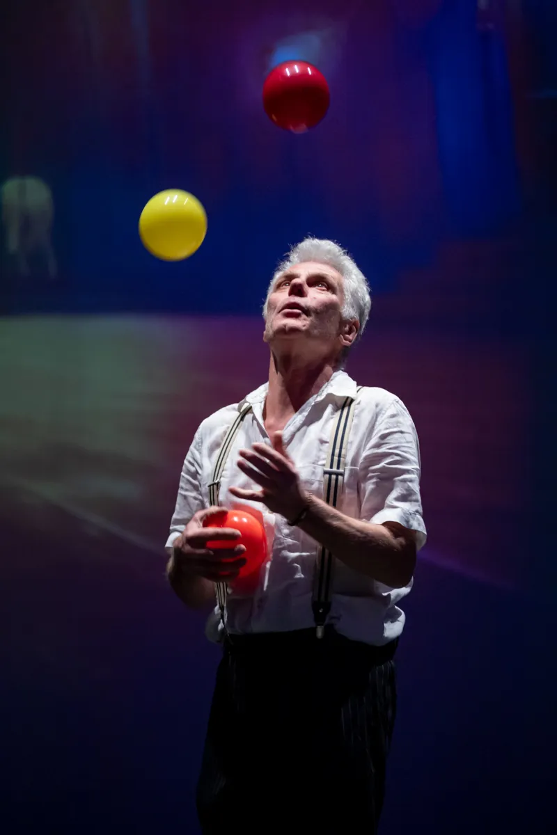 Ein älterer Mann mit weißen Haaren jongliert mit drei Bällen auf einer Bühne. Er trägt ein weißes Hemd mit Hosenträgern und eine dunkle Hose.