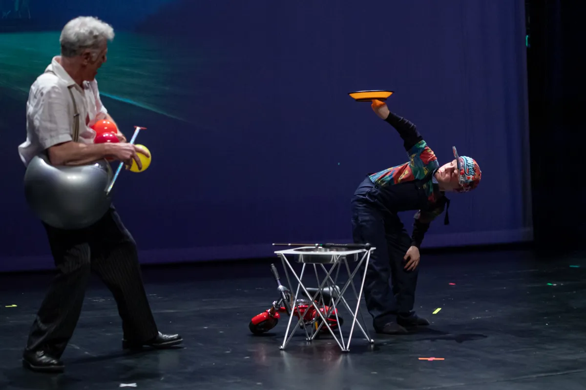 Zwei Clowns auf einer Bühne, einer jongliert mit einem Teller, während der andere einen roten Ball hält.