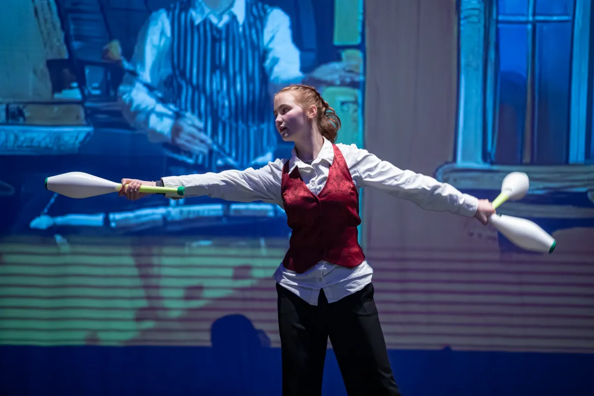 Junge Frau jongliert mit drei Keulen vor einer verschwommenen Kulisse mit einem Mann, der ein Instrument spielt.