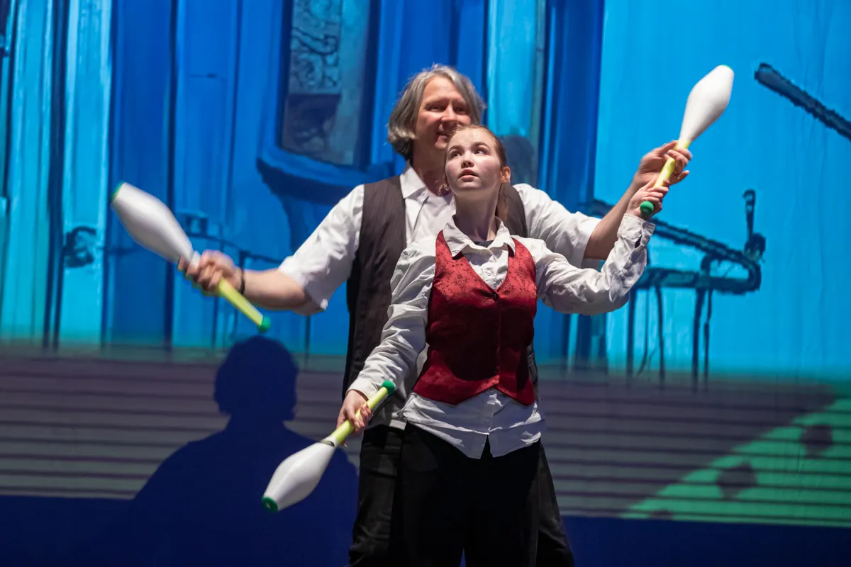 Zwei Artisten, eine Frau und ein Mann, jonglieren mit Keulen auf einer Bühne vor einer stilisierten Stadtkulisse.