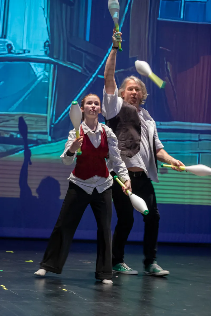 Zwei Jongleure, eine junge Frau und ein älterer Mann, werfen Keulen auf einer Bühne. Im Hintergrund eine stilisierte Stadtlandschaft in Blautönen.