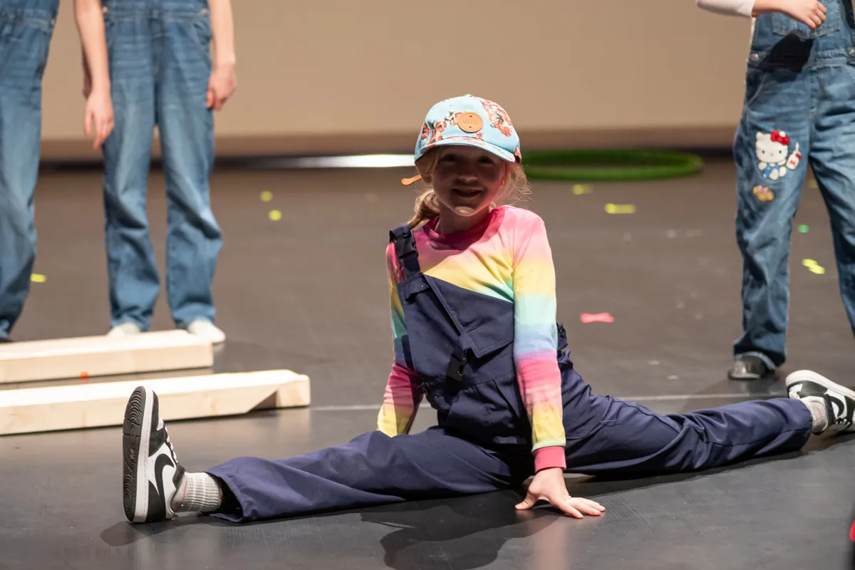 Ein Mädchen in einem bunten Regenbogenpullover und blauen Overalls sitzt auf der Bühne in Spagat und lächelt in die Kamera. Im Hintergrund stehen weitere Kinder in Jeans und Overalls.