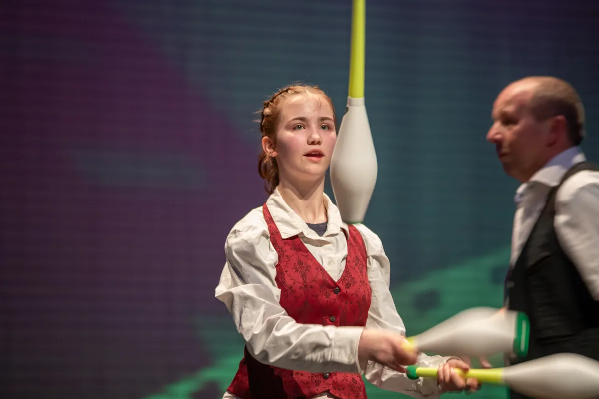 Ein junges Mädchen mit roten Haaren jongliert mit Keulen, während ein Mann im Hintergrund einen Stab hält. Die beiden treten auf einer Bühne vor einem violetten Hintergrund auf.