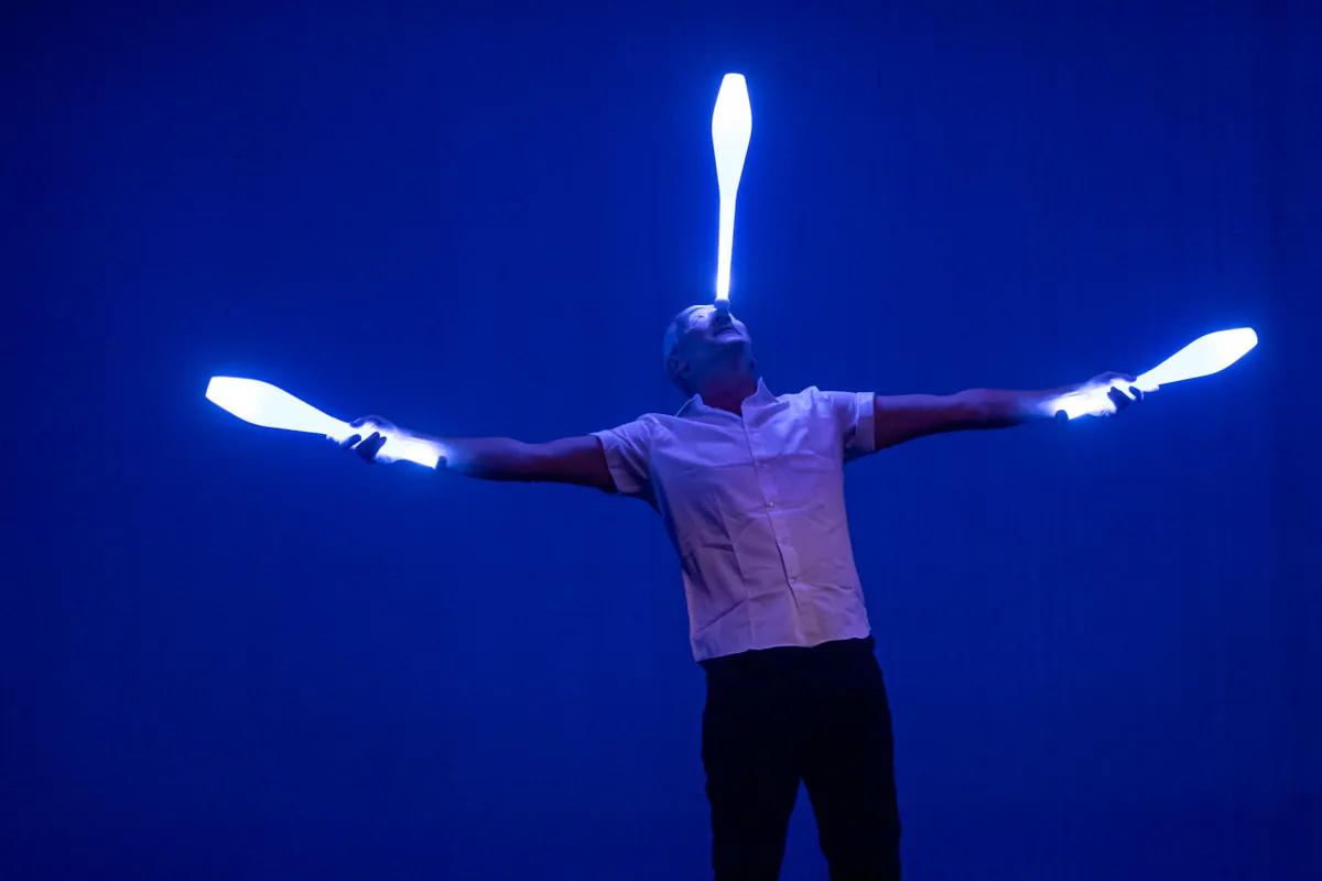 Ein Jongleur hält drei leuchtende Keulen in der Luft, eine auf dem Kopf und je eine in jeder Hand.