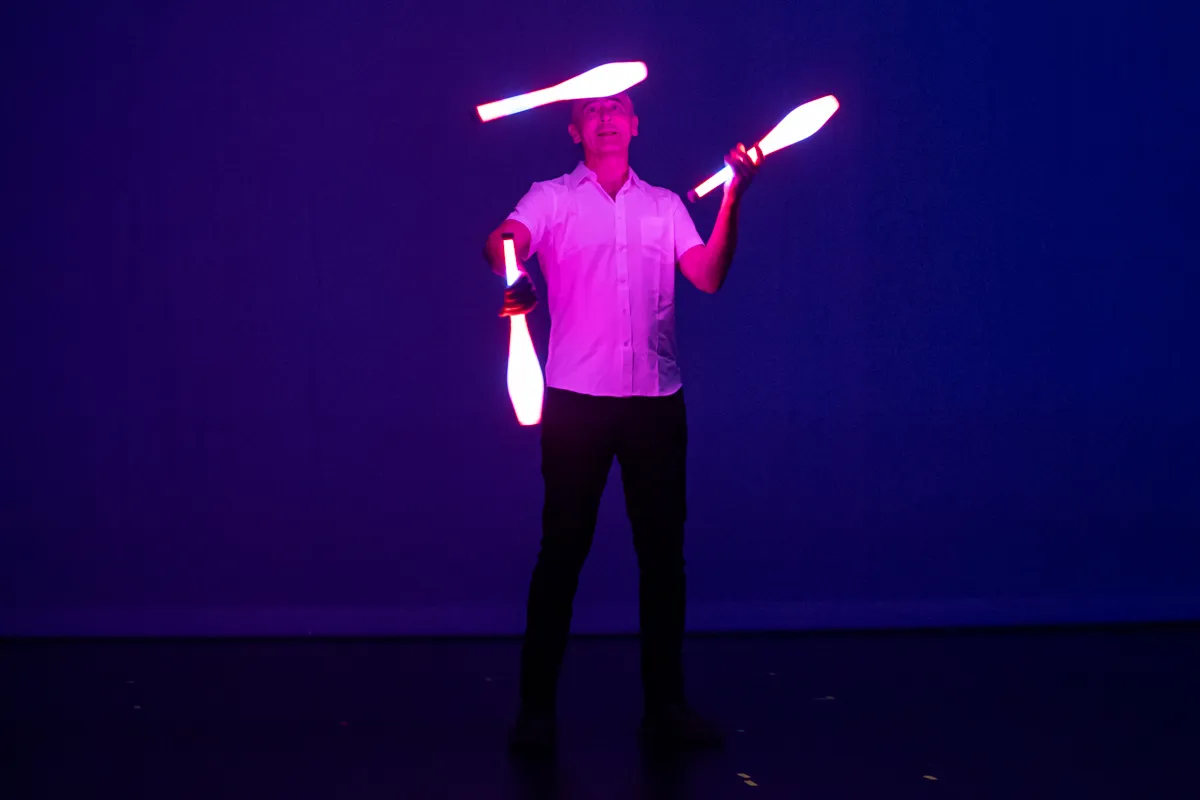 Ein Mann jongliert mit leuchtenden Keulen vor einem violetten Hintergrund. Er trägt ein weißes Hemd und dunkle Hosen.