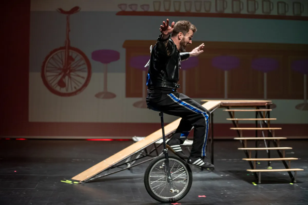 Ein Mann fährt auf einem Einrad über eine Rampe auf einer Bühne. Im Hintergrund sind weitere Requisiten und ein roter Vorhang zu sehen.