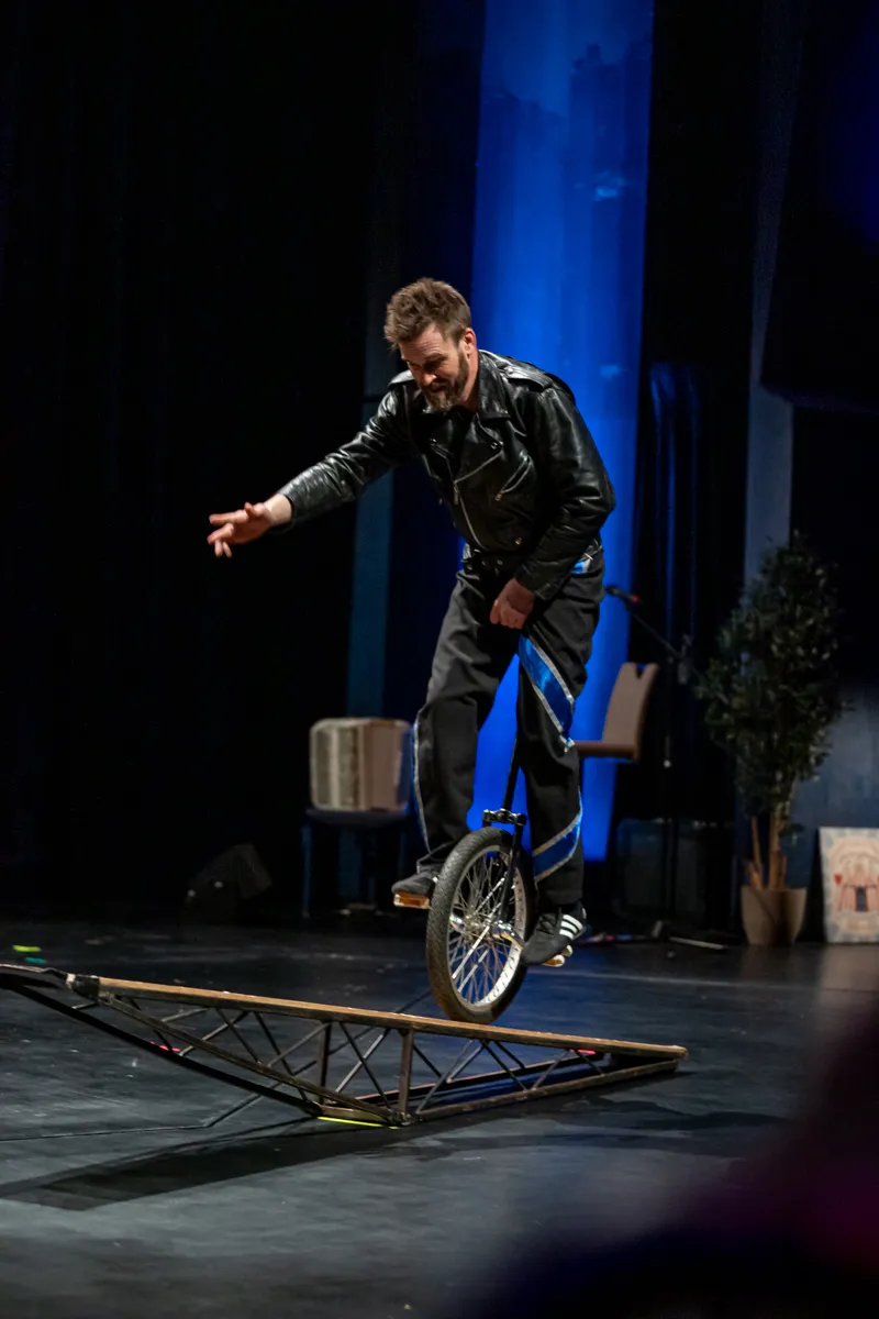 Ein Mann fährt auf einem Einrad über eine Wippe auf einer Bühne. Er trägt eine Lederjacke und dunkle Hosen.