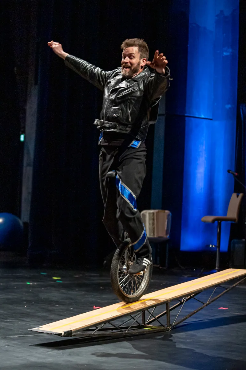 Ein Mann fährt auf einem Einrad über eine schräge Rampe, die Arme sind zum Balancieren ausgebreitet. Im Hintergrund eine Bühne mit blauer Beleuchtung und Stühlen.