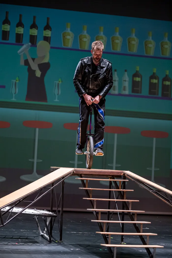 Ein Mann fährt auf einem Einrad auf einer schmalen Holzkonstruktion über einer Treppe, während im Hintergrund eine stilisierte Bar mit Flaschen und einer Bedienenden zu sehen ist.