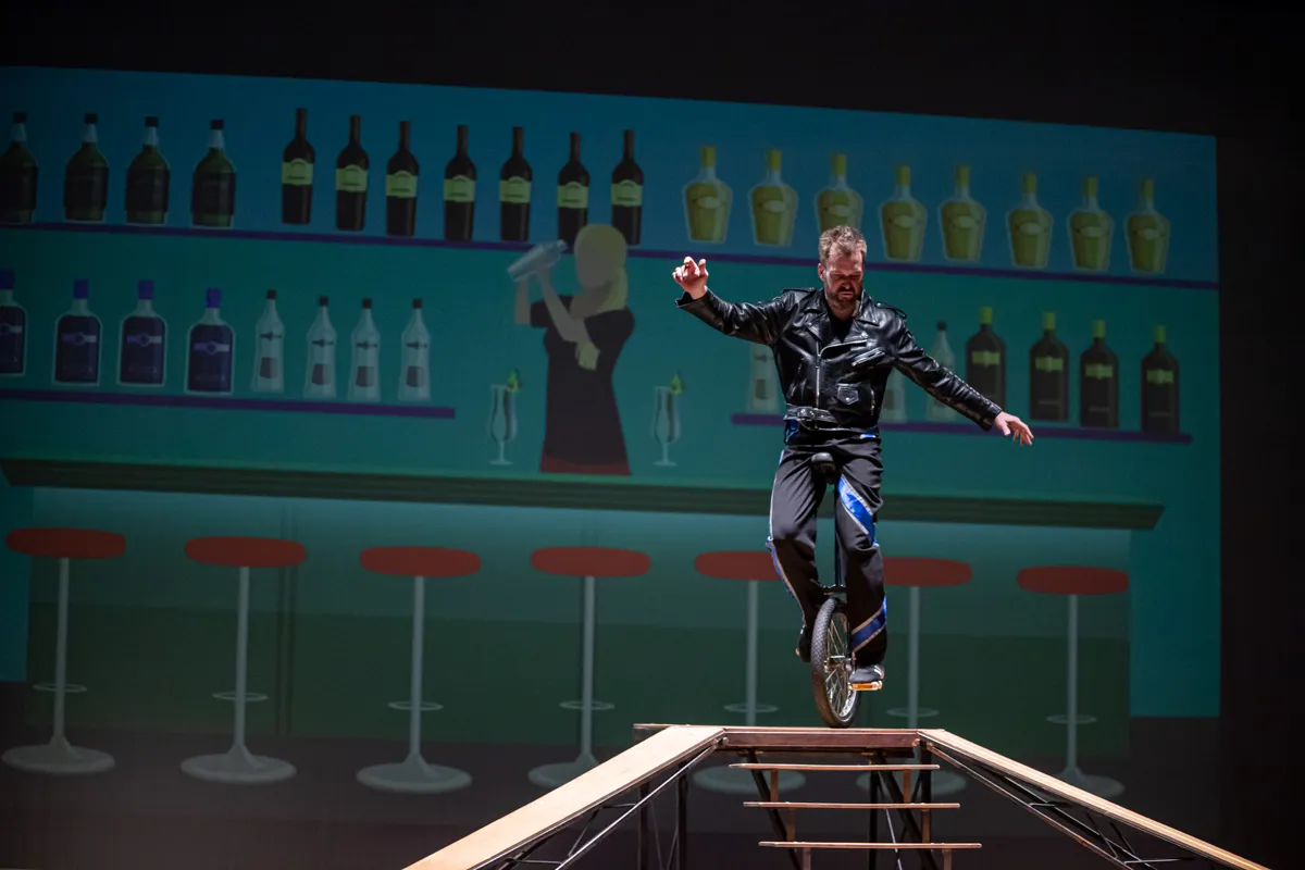 Ein Mann fährt auf einem Einrad auf einem schmalen Balken, während im Hintergrund eine stilisierte Bar mit Flaschen und Barhocker zu sehen ist.