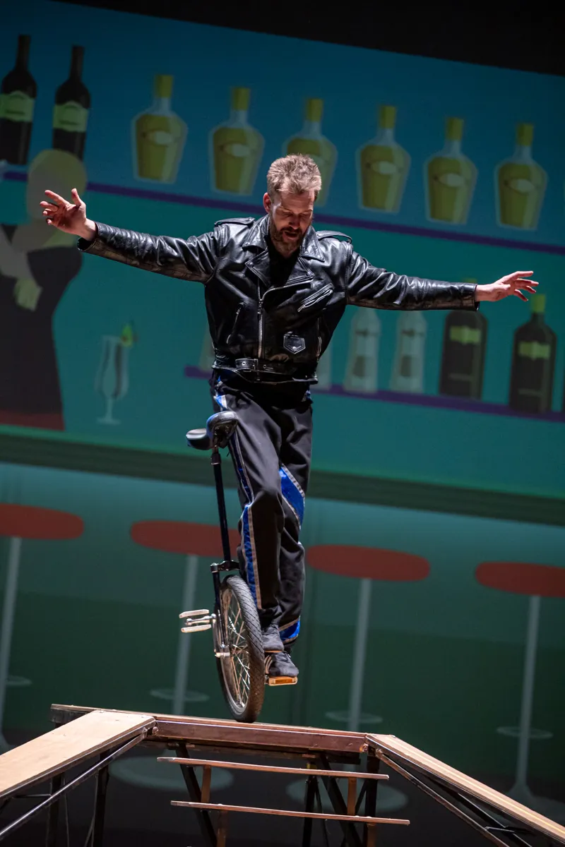 Ein Mann mit Bart fährt auf einem Einrad auf einem schmalen Balken, die Arme sind zum Ausgleich ausgebreitet. Im Hintergrund eine stilisierte Bar mit Flaschen und Barhocker.