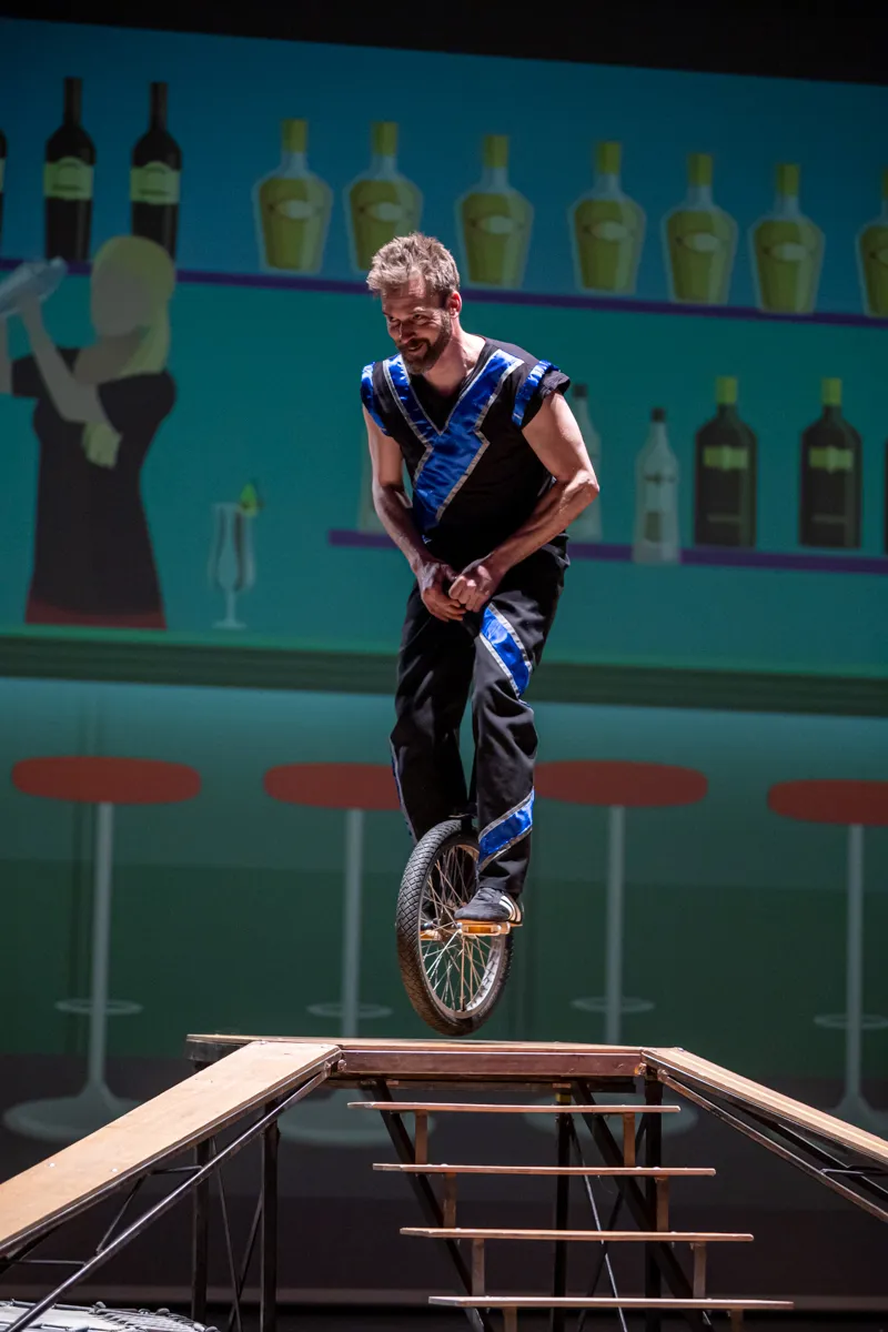 Ein Mann fährt auf einem Einrad auf einer schrägen Rampe vor einer Kulisse, die eine Bar mit Flaschen und einem stilisierten Kellner darstellt.