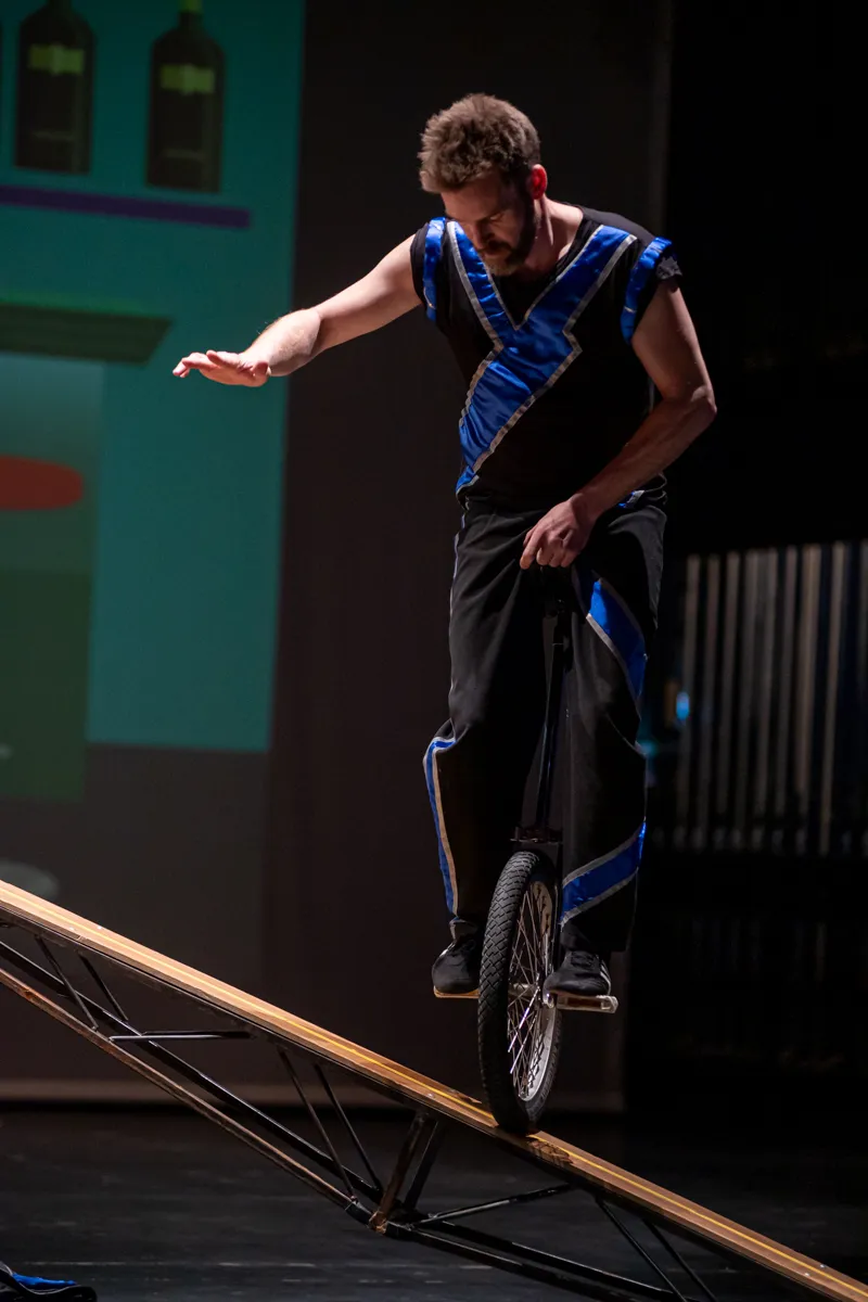 Ein Mann mit Bart fährt auf einem Einrad über eine schräge Rampe, während er die Balance hält. Er trägt ein blau-schwarzes Outfit.