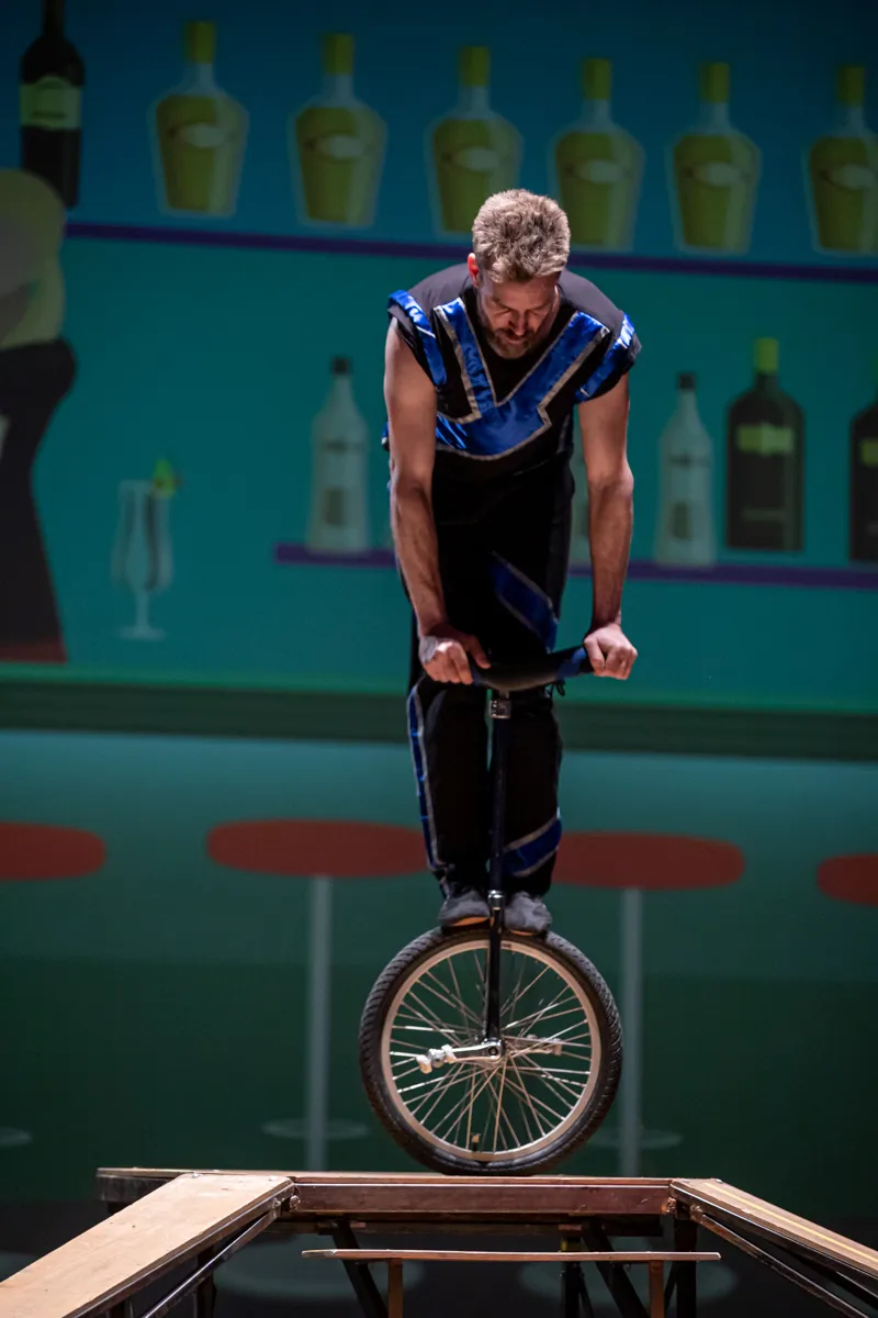 Ein Mann fährt auf einem Einrad auf einer niedrigen Holzplattform, während er sich vornüberbeugt. Im Hintergrund stehen Flaschen und ein Weinglas auf einem Regal.
