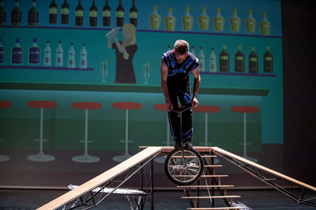 Ein Mann fährt auf einem Einrad auf einem schmalen Balken, während im Hintergrund eine stilisierte Bar mit Flaschen und Barhocker zu sehen ist.