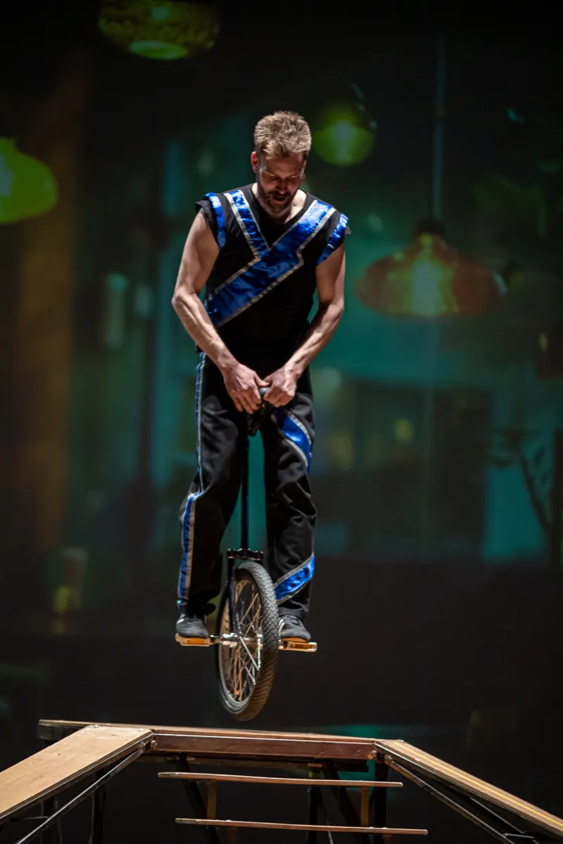Ein Mann fährt auf einem Einrad auf einer schmalen Holzplanke, während er sich mit dem Kopf nach unten beugt.