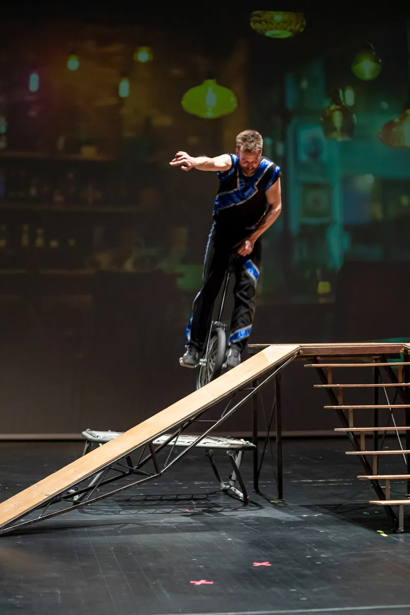 Ein Mann fährt auf einem Einrad auf einer schiefen Rampe über einer Trampolin-artigen Fläche, während er die Balance hält.