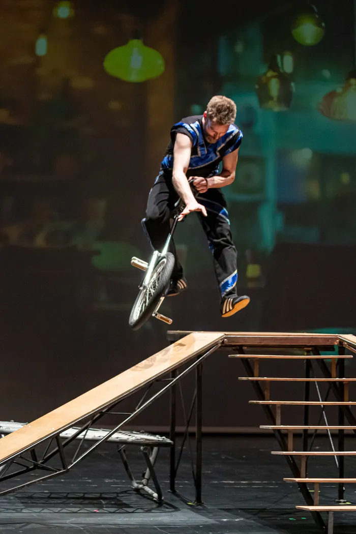 Ein Mann springt mit einem Einrad über eine Rampe und eine Treppe auf einer Bühne. Im Hintergrund sind verschwommene Lichter und Formen zu sehen.