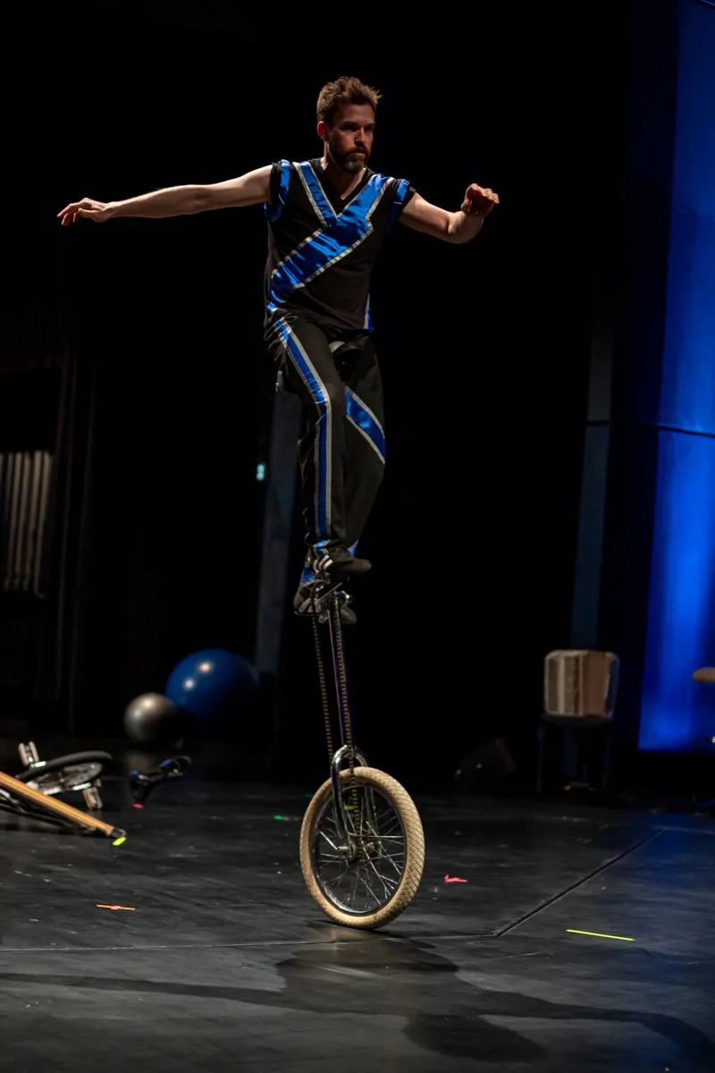 Ein Mann fährt auf einem Einrad auf einer Bühne, die Arme sind zum Ausgleich ausstreckend. Im Hintergrund sind Requisiten und blaues Licht zu sehen.
