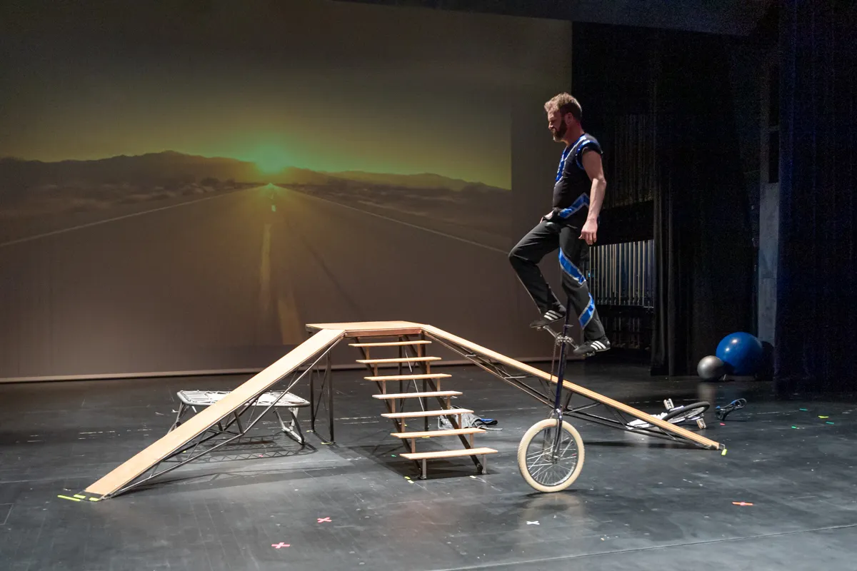 Ein Mann fährt auf einem Einrad über eine Rampe und eine kurze Treppe auf einer Bühne vor einer Leinwand mit einer Landschaftsaufnahme.
