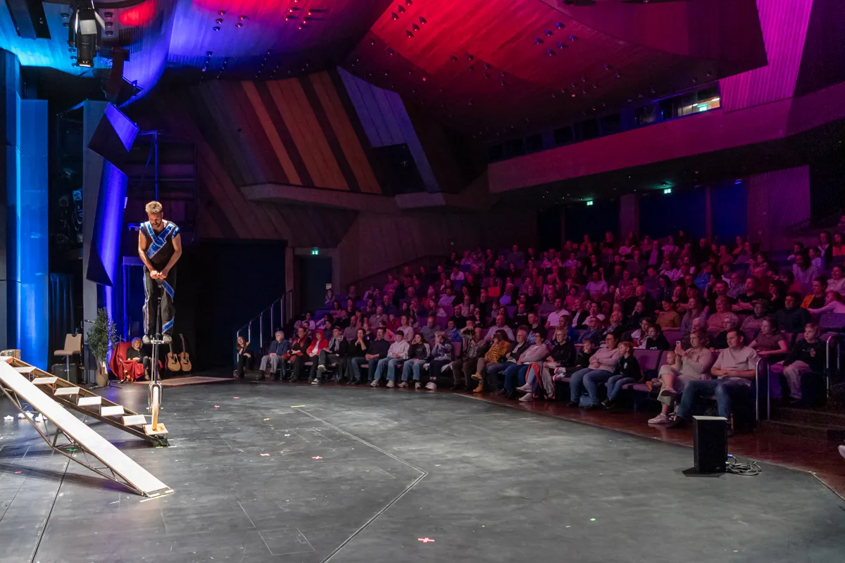 Ein Künstler auf Stelzen balanciert auf einer Rampe vor einem voll besetzten Saal. Das Publikum schaut auf die Bühne, die in rotes und blaues Licht getaucht ist.