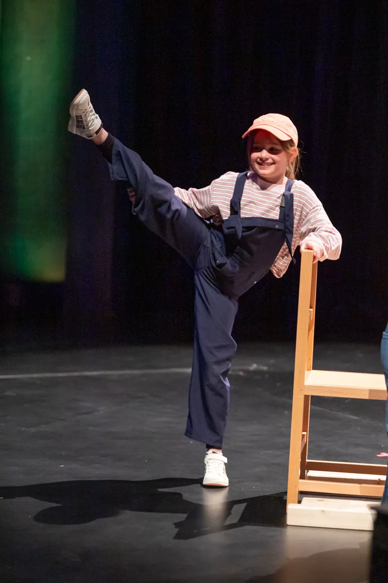 Ein Kind in einem Overall und einer Kappe steht auf einer Bühne und führt eine Tanzbewegung auf, während es sich an einem Stuhl festhält.