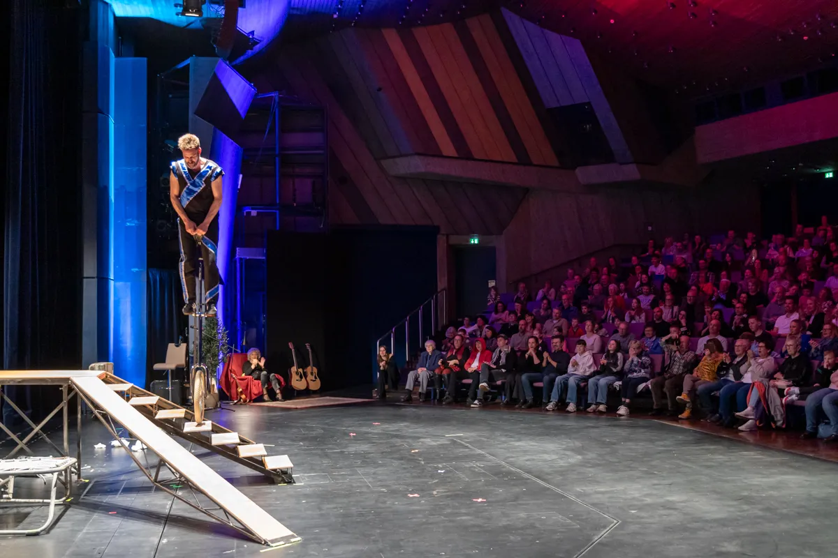 Ein Akrobat balanciert auf Stelzen auf einer schiefen Rampe vor einem Publikum in einem Theater. Im Hintergrund eine Bühne mit Gitarren und rotem Licht.