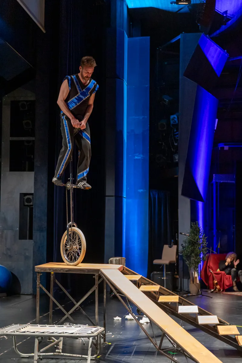 Ein Mann fährt auf einem Einrad über eine Rampe auf einer Bühne. Im Hintergrund sind blaue Säulen und ein abstraktes Bühnenbild zu sehen.