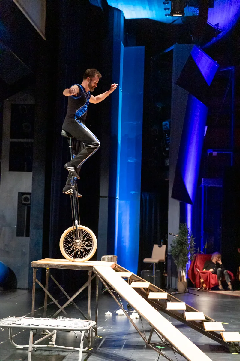 Ein Mann fährt auf einem Einrad über eine Rampe auf einer Bühne. Im Hintergrund eine blaue Kulisse und ein paar Zuschauer.