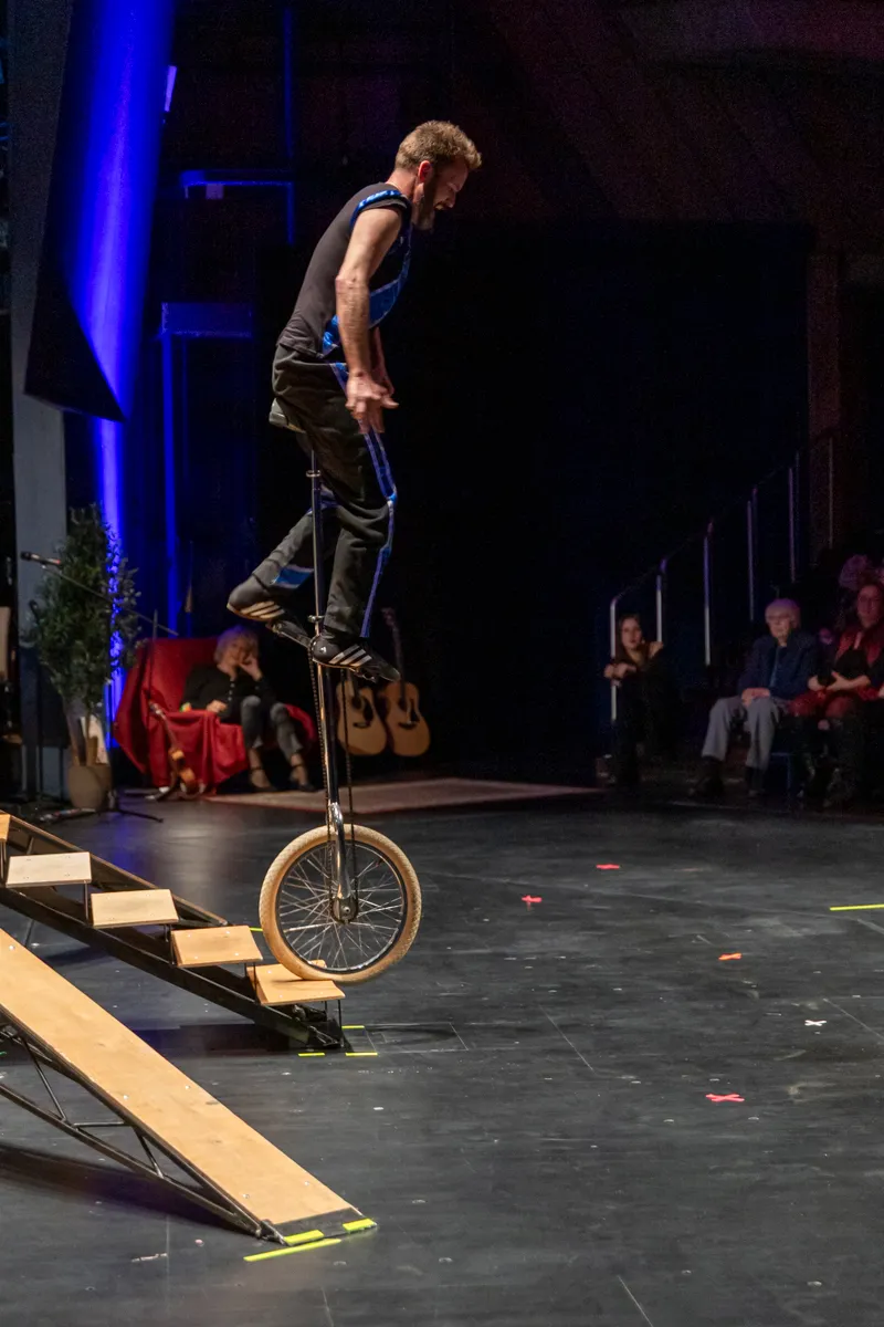 Ein Mann fährt auf einem Einrad über eine Rampe auf einer Bühne, während im Hintergrund Zuschauer sitzen.