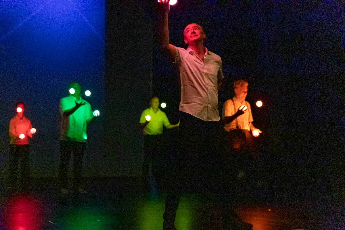 Ein Mann in weißem Hemd hält einen leuchtenden Ball über seinem Kopf, umgeben von weiteren Personen mit leuchtenden Kugeln in den Händen auf einer dunklen Bühne.