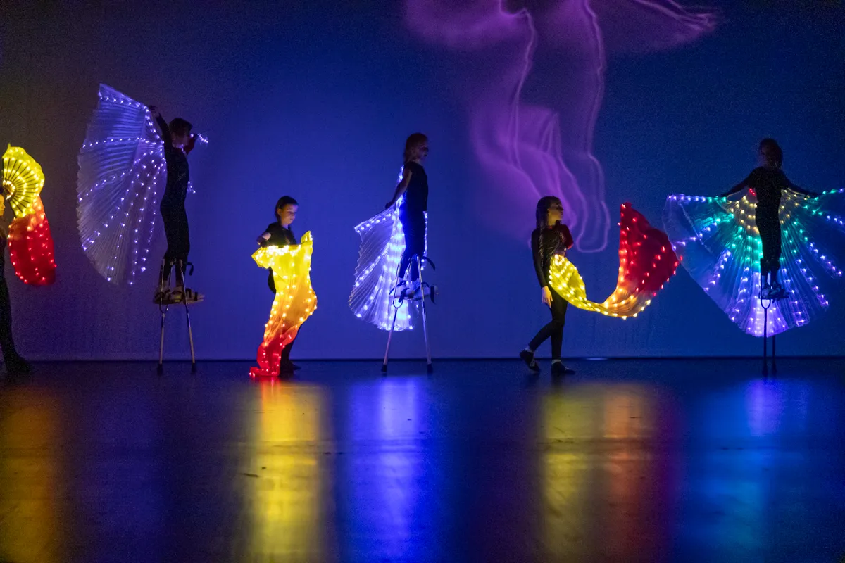 Fünf Kinder und Jugendliche in fantasievollen Kostümen mit leuchtenden Flügeln treten auf einer Bühne auf. Im Hintergrund ist eine abstrakte Projektion zu sehen.