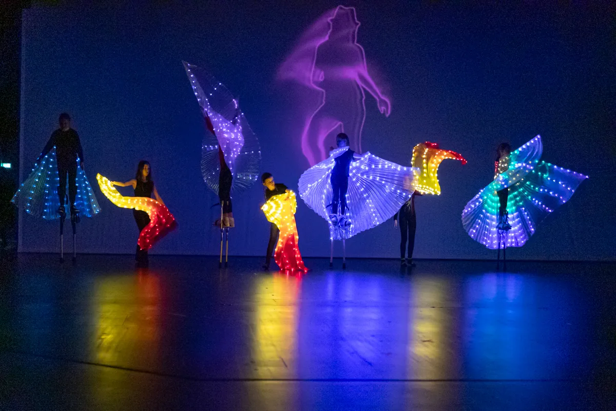 Fünf Darsteller mit leuchtenden Flügeln in verschiedenen Farben stehen auf einer Bühne vor einer blauen Wand mit einer silhouettenartigen Projektion einer Frau. Buntes Licht beleuchtet den Bühnenboden.