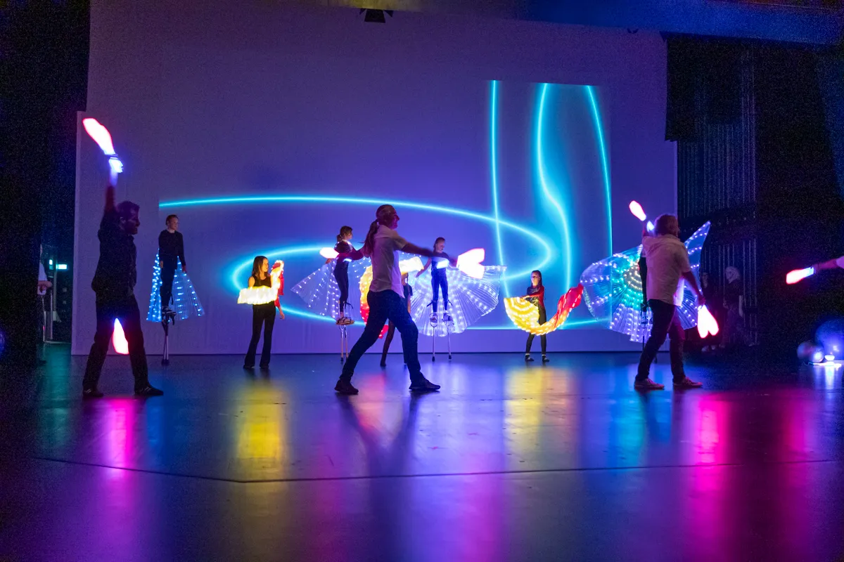 Mehrere Darsteller in farbenfrohen Kostümen und mit leuchtenden Requisiten treten auf einer Bühne vor einer blauen Lichtinstallation auf. Die Performance ist Teil der Premiere der Show „Kaskadia“.