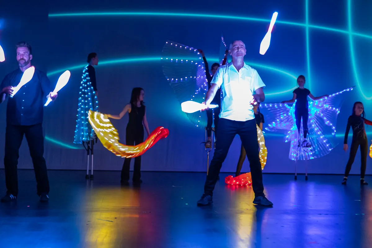 Ein Mann jongliert mit leuchtenden Keulen auf einer Bühne, umgeben von weiteren Darstellern mit leuchtenden Kostümen und Accessoires.