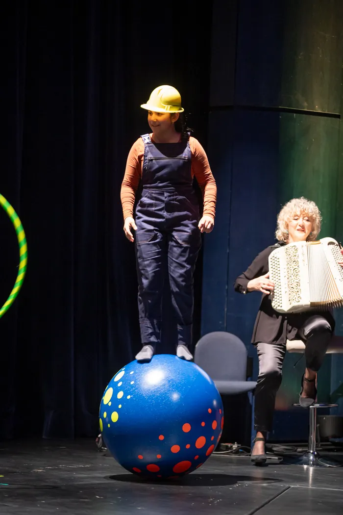 Eine Person in Latzhose und gelbem Helm steht auf einem blauen Ball auf einer Bühne, während eine Frau im Hintergrund ein Akkordeon spielt.