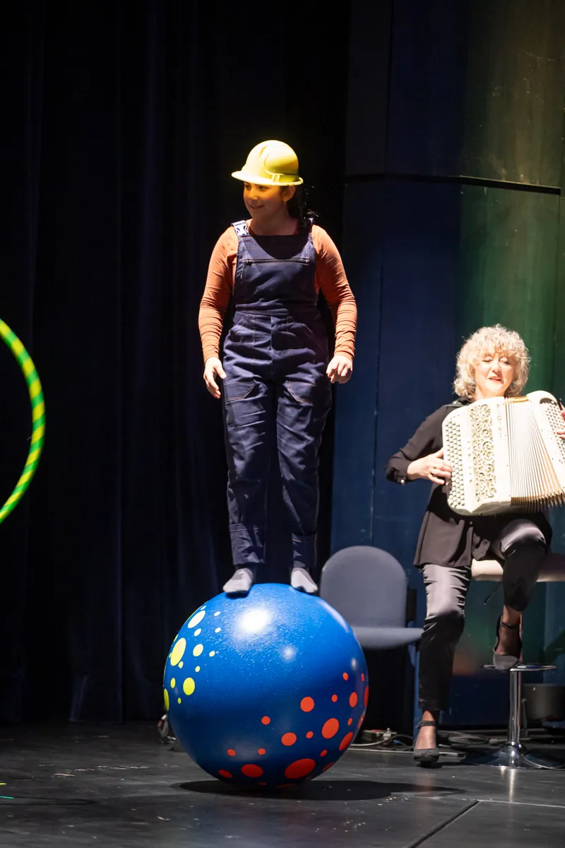 Eine Person in Latzhose und gelbem Helm steht auf einem blauen Ball auf einer Bühne, während eine Frau im Hintergrund ein Akkordeon spielt.