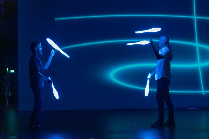 Zwei Artisten jonglieren mit leuchtenden Keulen vor einer blauen Wand mit Lichtlinien auf einer Bühne.