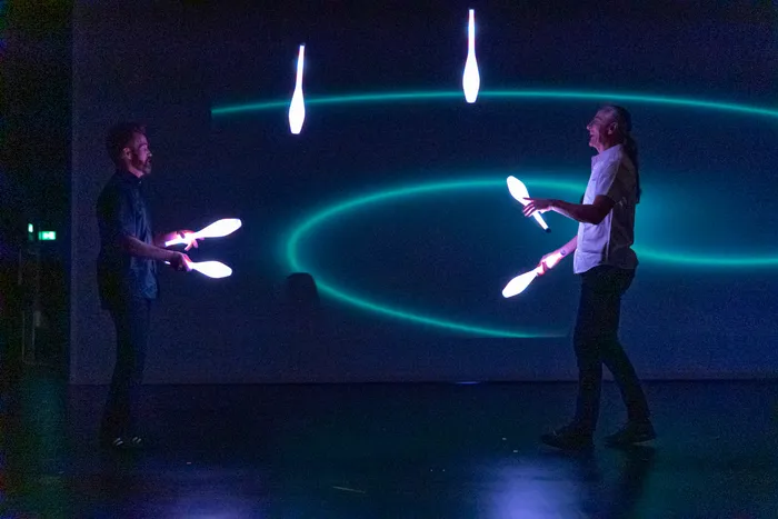Zwei Artisten jonglieren mit leuchtenden Objekten vor einem dunklen Hintergrund mit blauen Lichtlinien.
