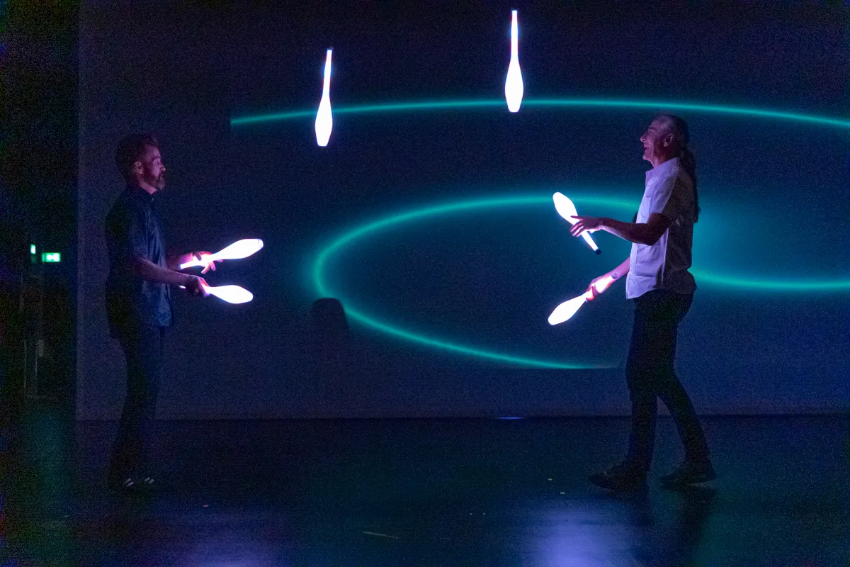 Zwei Artisten jonglieren mit leuchtenden Objekten vor einem dunklen Hintergrund mit blauen Lichtlinien.