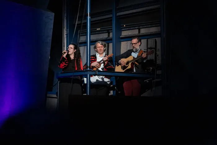 Drei Musiker spielen auf einer erhöhten Bühne: eine Frau singt in ein Mikrofon, während zwei Männer Gitarre und ein weiteres Saiteninstrument spielen.